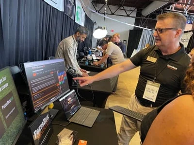 Greg Gibbs pointing to the Press Box Toolkit app on a monitor while talking with Demo Day attendees